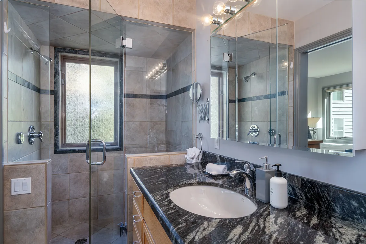 A modern bathroom featuring a glass shower enclosure, granite countertop, and mirrored cabinets. The space is bright and tranquil, ideal for both quick morning routines and relaxing self-care rituals after a day in the mountains.