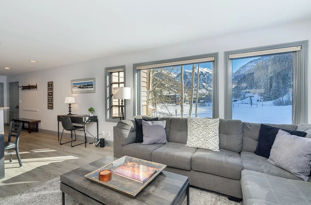 A tranquil living space with a comfortable sectional sofa, a wooden coffee table, and expansive windows that provide unobstructed mountain views. The ambiance is peaceful, flooded with natural light, and perfect for relaxation or gathering with family and friends.