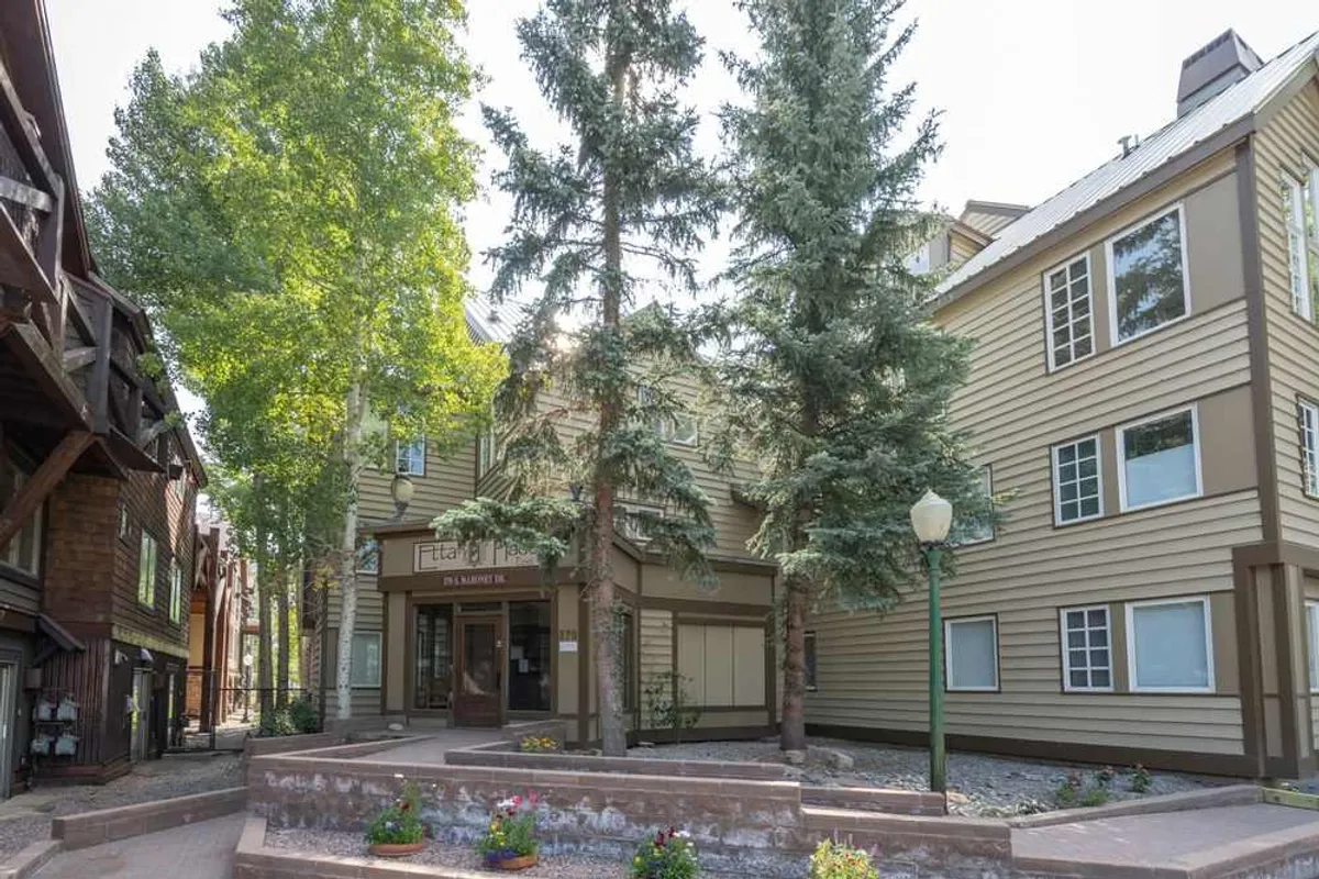 A welcoming photo of the building's entrance, framed by tall trees and vibrant flower beds. The inviting path and exterior details evoke a sense of arrival and easy outdoor access.