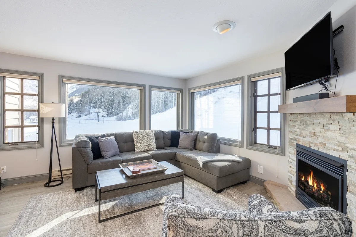 Inside, a cozy sofa faces a flickering fireplace, with windows framing a glimpse of snow-capped peaks. The inviting living area suggests relaxation and connection, paired with the beauty visible just beyond the glass.