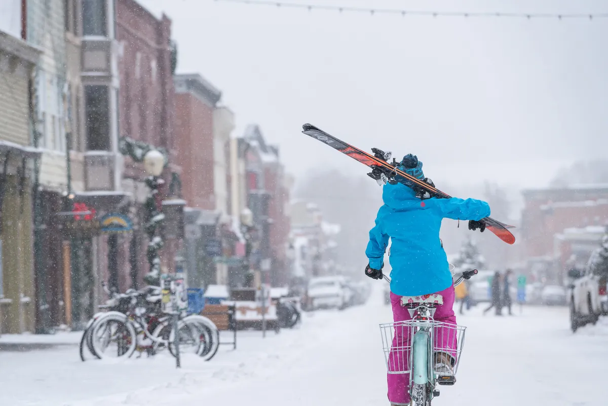 A person rides a bicycle with skis slung over their shoulder along a snow-covered street surrounded by historic buildings. The scene captures Telluride’s unique blend of outdoor adventure and small-town charm, inviting group members to discover local attractions and embrace the wintery ambiance.