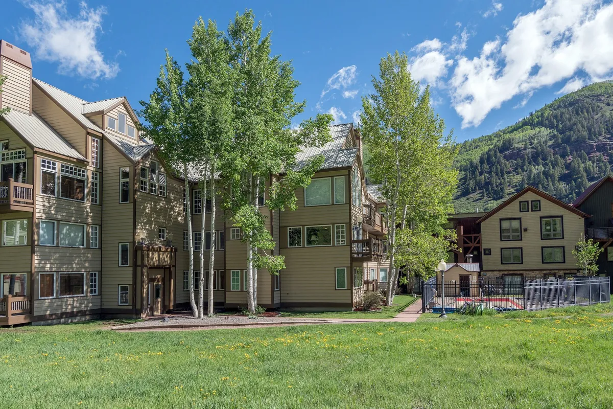 The exterior of an apartment building framed by lush trees, with mountains rising in the background and a pool area in the foreground. The scene suggests both relaxation and immediate access to outdoor activities, with clear skies and inviting communal amenities.