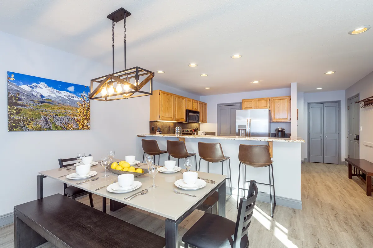 This image displays a welcoming dining area with a set table, a kitchen counter with bar stools, and mountain-themed artwork. The space feels homey and is suited for family meals or gatherings.