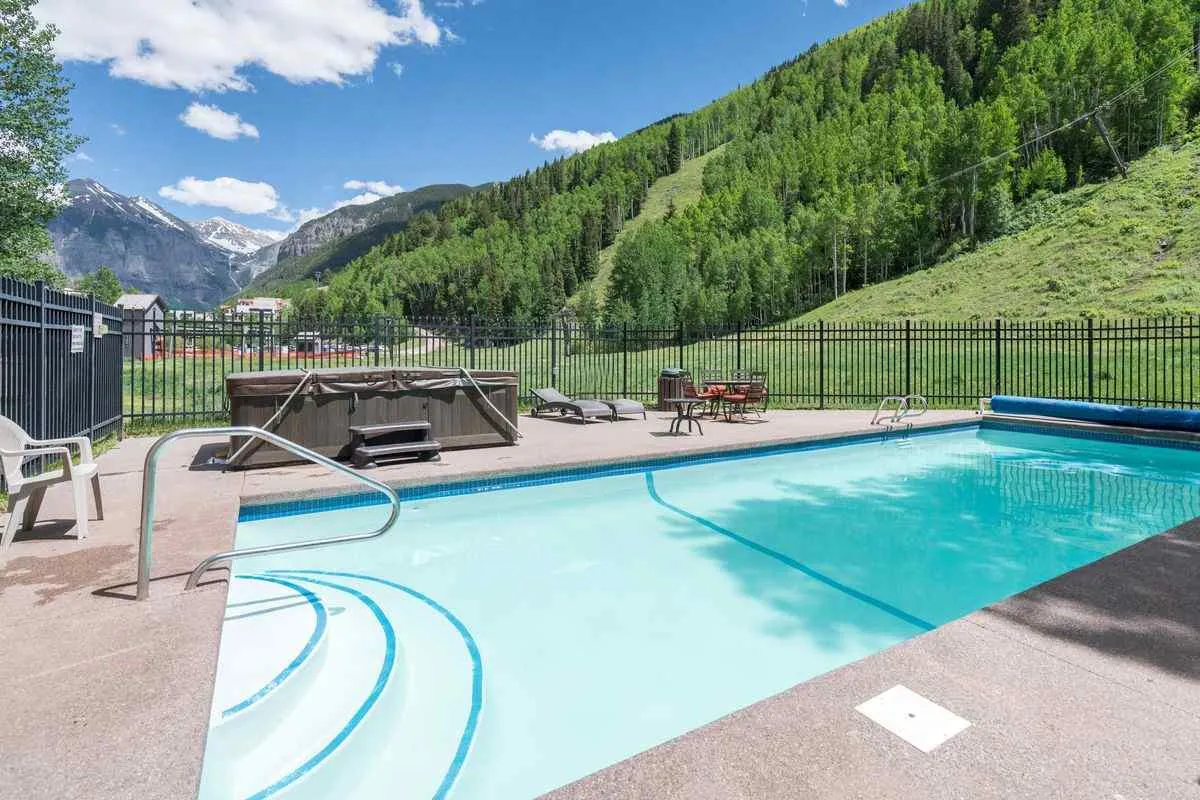 An inviting exterior scene with a swimming pool and hot tub set against a mountain backdrop and lush greenery. The space is designed for both relaxation and fun, highlighting shared amenities available to guests, including soaking after skiing or swimming in summer.