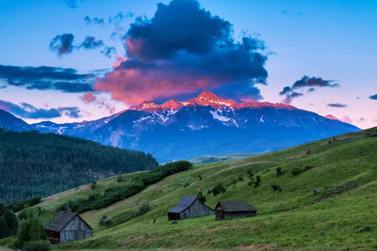 This image showcases a sweeping view of snow-capped mountains at sunset, with dramatic clouds, lush green hills, and rustic wooden cabins nestled below. The tranquil setting invites viewers to imagine nature walks, breathtaking sunsets, and serene, intimate moments.