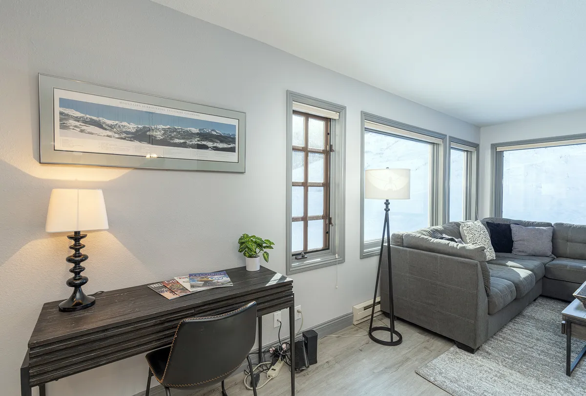 A calm, comfortable interior scene with a desk and lamp, sofa, wall artwork, and large windows letting in natural light. The setup suggests both relaxation and productivity, suitable for varied guest needs.