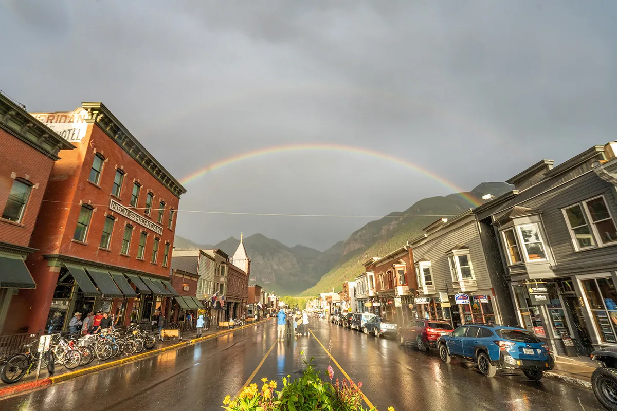 A lively exterior street scene featuring historic brick buildings, a rainbow arching across the sky, and bicycles parked along wet pavement. The area exudes small-town charm with vibrant local life and accessible shops.