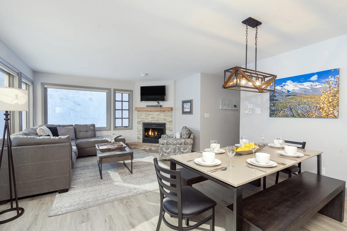 A warm, open living and dining space featuring a cozy gas fireplace, a plush sofa, stylish artwork, and a dining table set against large windows that showcase mountain views. The atmosphere is inviting, with natural light streaming in and a layout that encourages connection.
