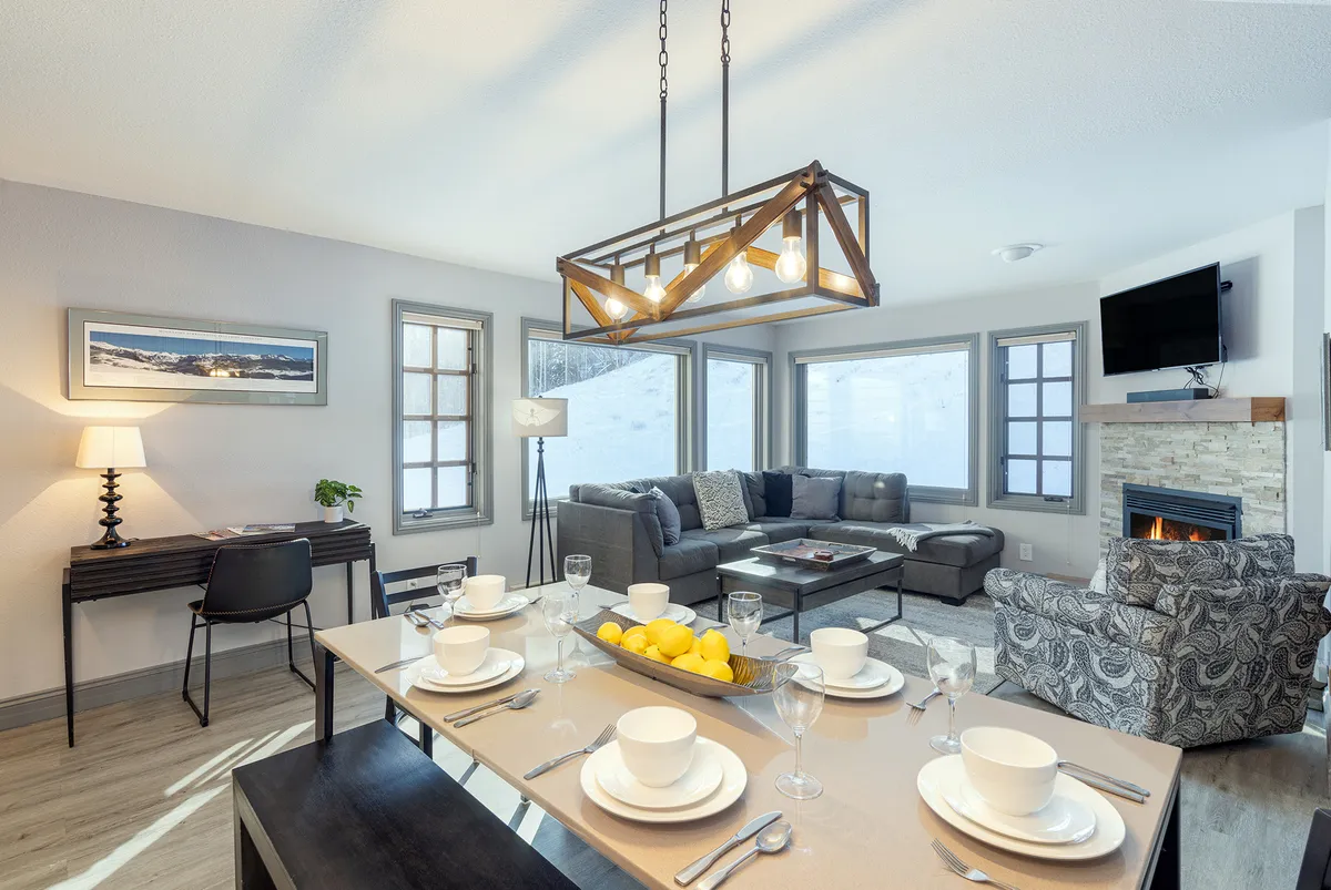 The image showcases a warmly lit, inviting open-plan living space featuring a dining table elegantly set for a meal, a plush seating area arranged around a glowing fireplace, and large windows hinting at mountain views. The setup encourages memorable gatherings, cozy evenings, and shared moments with loved ones.