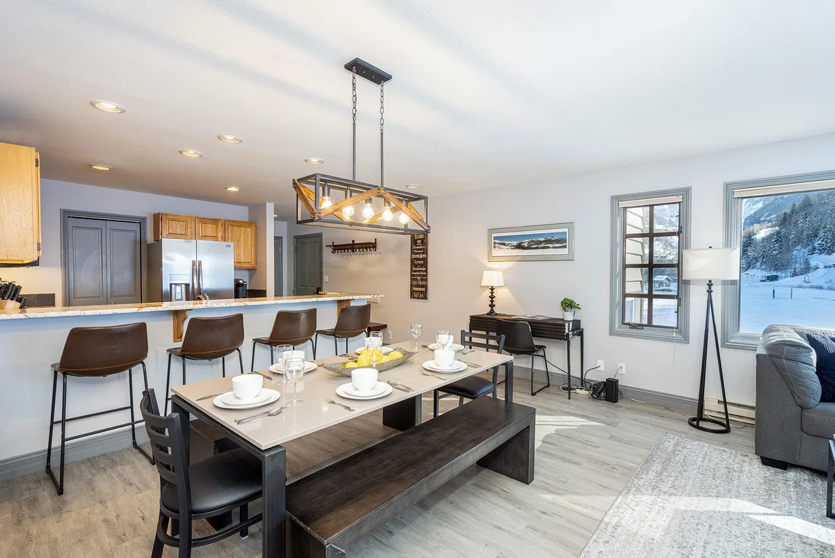 A seamless open-plan space highlighting a dining table with place settings, a breakfast bar with stools, and a living area with a plush sofa. Large windows frame stunning outdoor views, bathing the space in natural light and reinforcing the sense of togetherness.