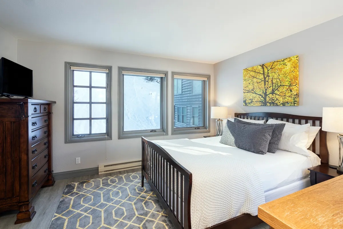 A serene bedroom featuring a comfortable bed with decorative pillows, large windows that let in ample natural light, tasteful artwork above the dresser, and a soft rug on the floor. The setting is peaceful and inviting, suggesting a restful retreat ideal for privacy and relaxation.
