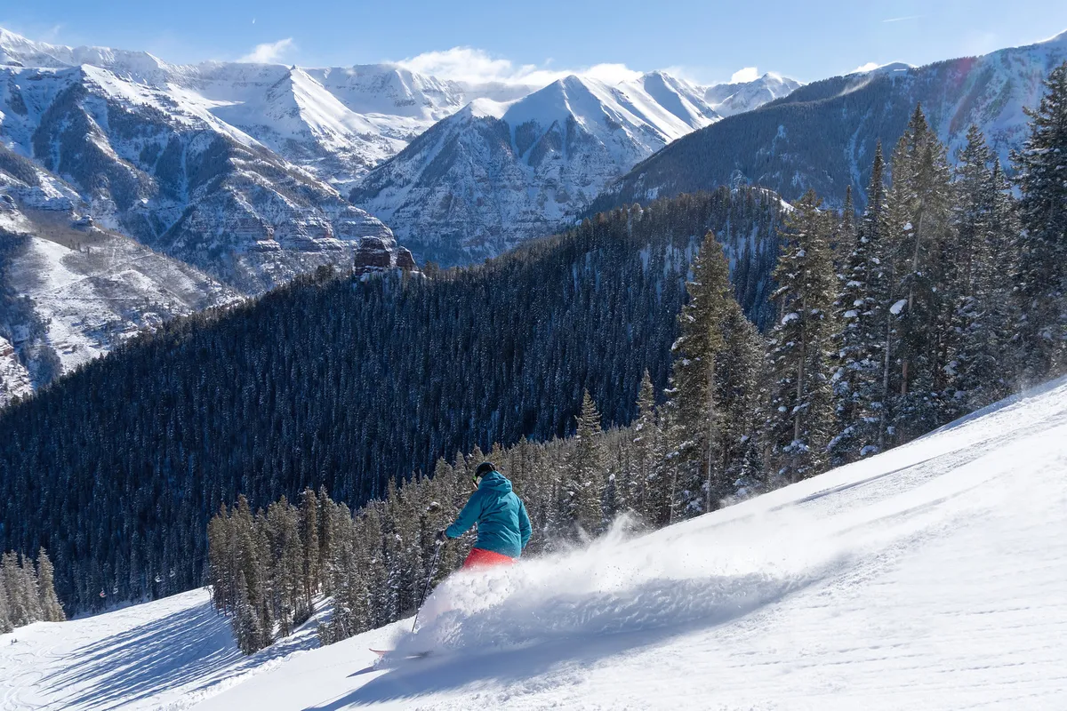 A skier gracefully navigates a powdery slope surrounded by snow-covered mountains and dense pine trees. The expansive landscape captures the thrill of downhill skiing with crisp white snow, a sweeping mountain backdrop, and the inviting allure of adventure on the slopes.