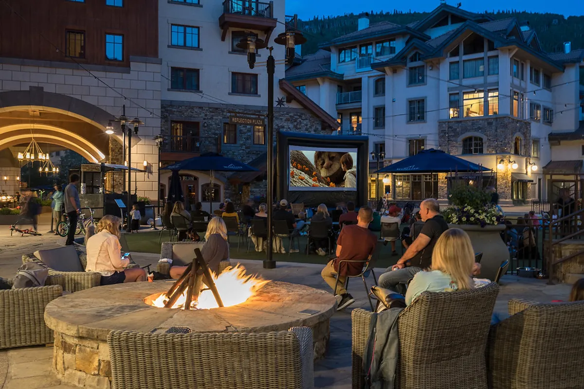 An inviting outdoor communal area featuring a glowing fire pit, comfortable seating, and an outdoor movie screen, set beneath the open sky. The setup evokes a relaxed and communal mood, perfect for group gatherings and evening relaxation after a day of adventure.