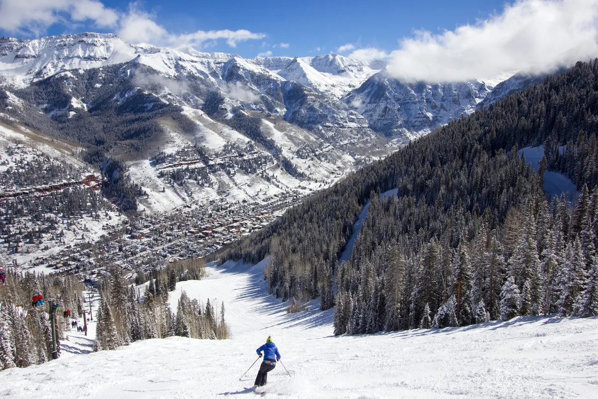 A skier descends a snowy mountain slope with rugged peaks and dense evergreens in the distance. Below, the charming town of Telluride nestles in the valley, offering a panoramic view that ties together mountain adventure and local exploration.