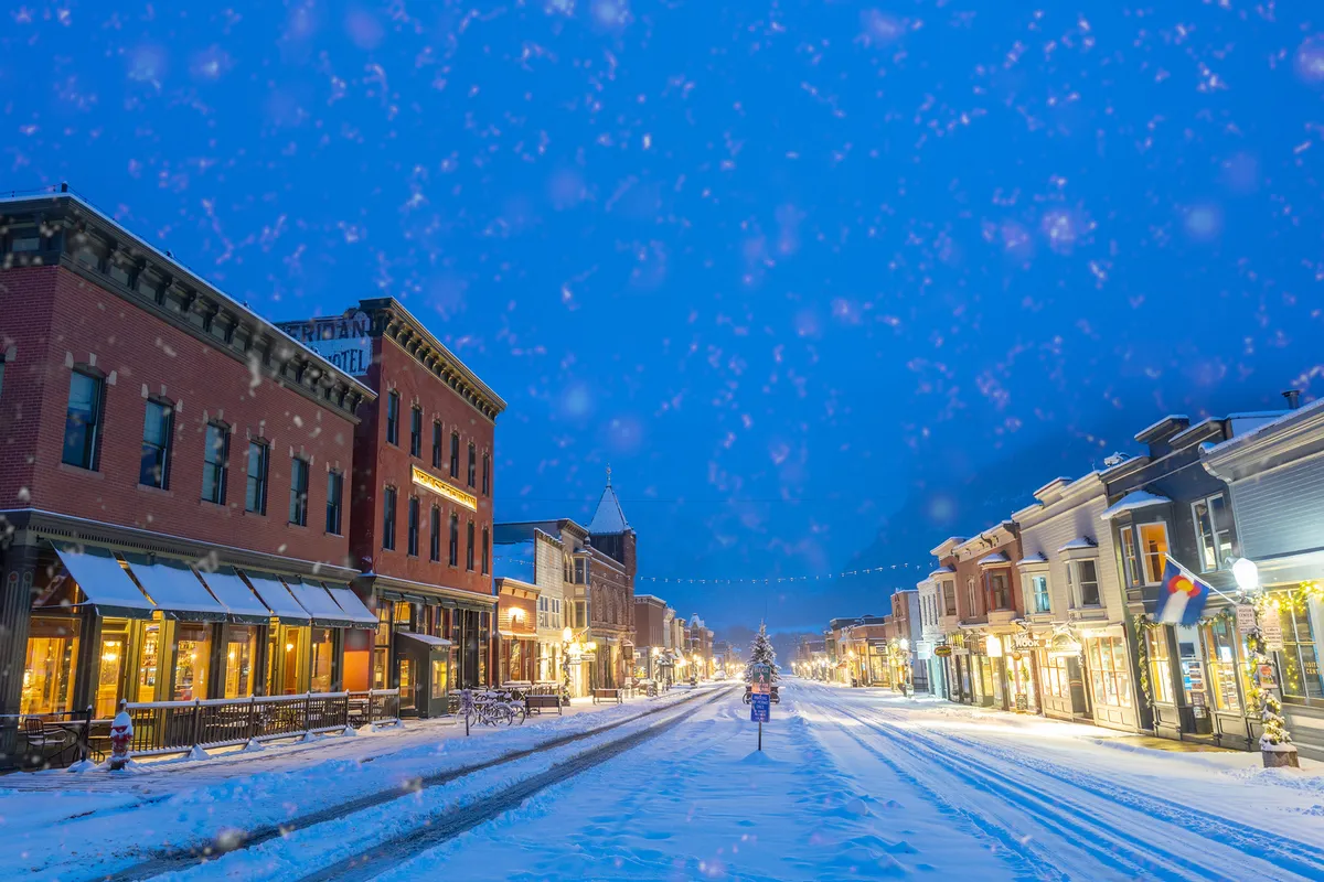 A snow-covered street lined with charming decorated storefronts, festive lights, and a Christmas tree visible in the distance. The image evokes a sense of holiday spirit, community, and the vibrant atmosphere of Telluride’s main street, with inviting pathways for strolling and shopping.